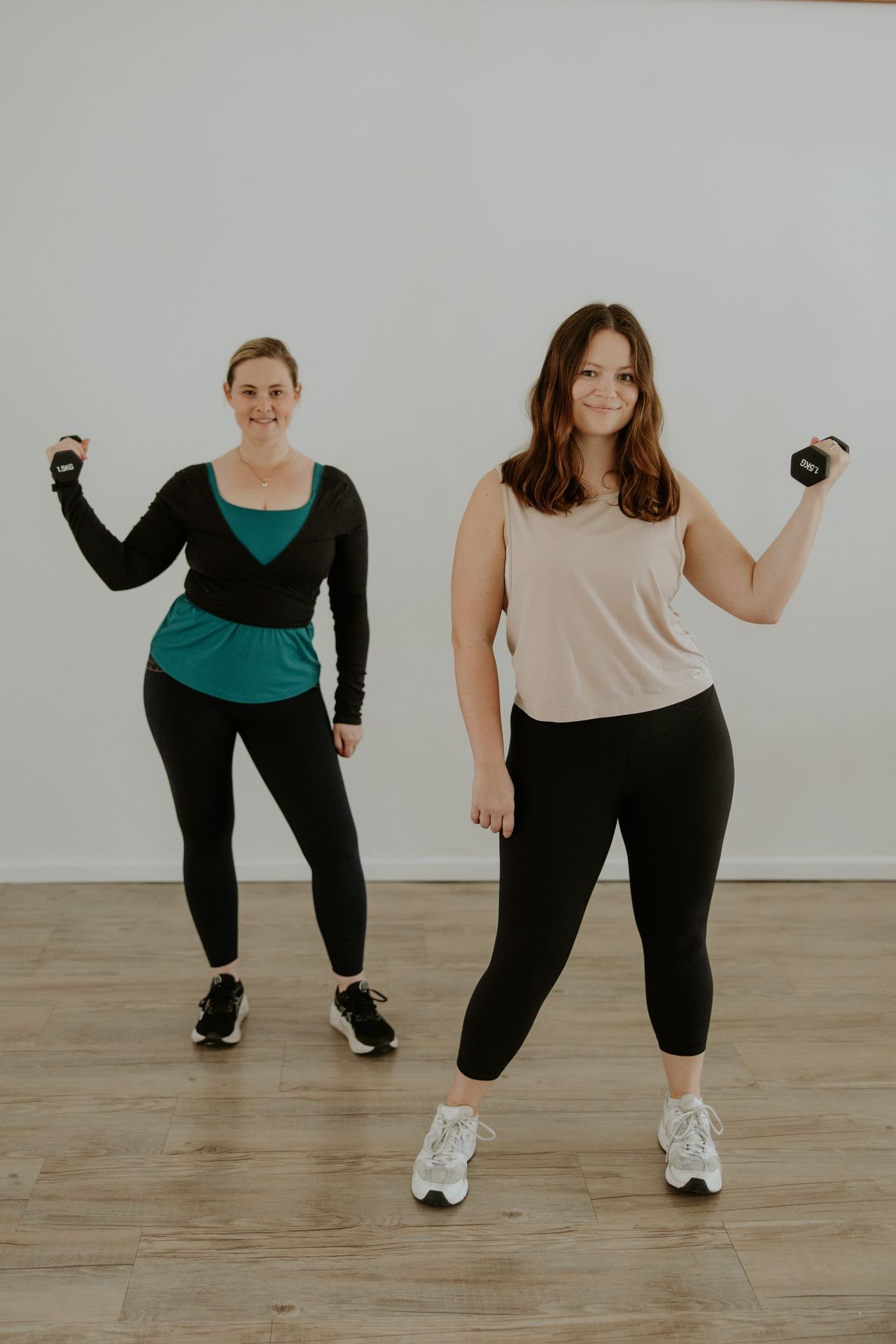 Two women in workout clothes lifting small dumbbells while standing on a wooden floor.