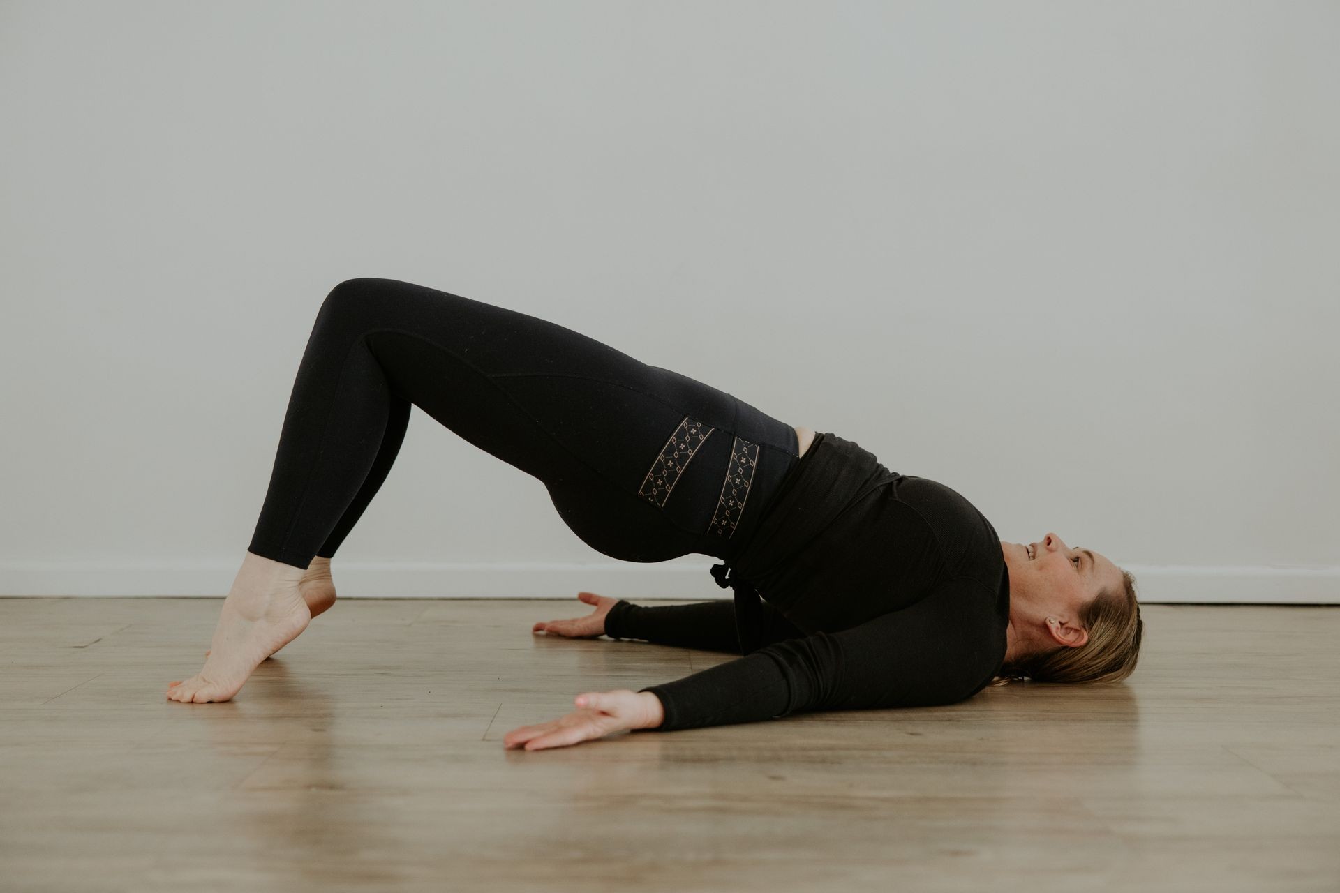 Person wearing black workout clothes performs a bridge pose on a wooden floor.