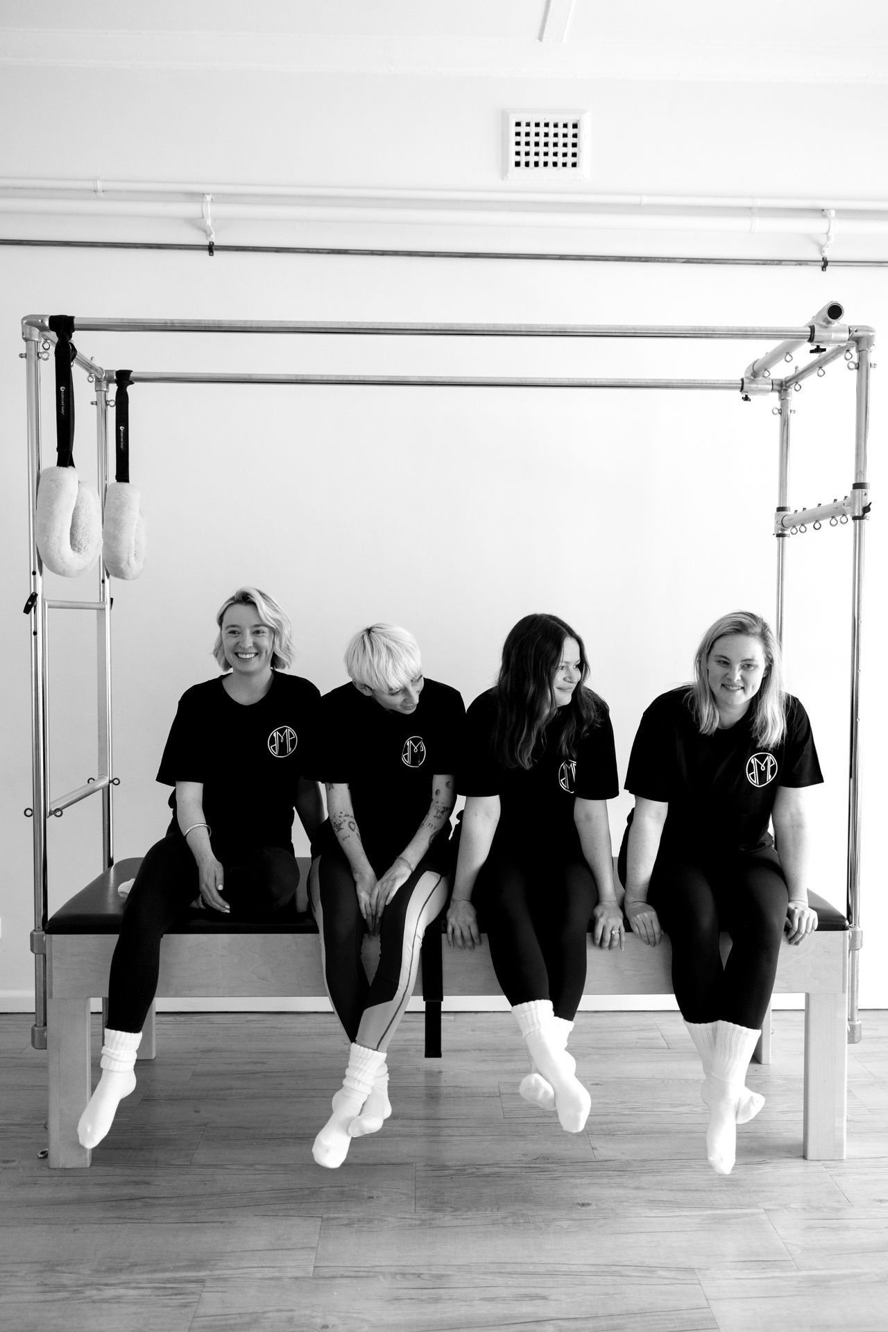 Four people wearing matching athletic attire sit on a piece of exercise equipment in a minimalist gym setting.